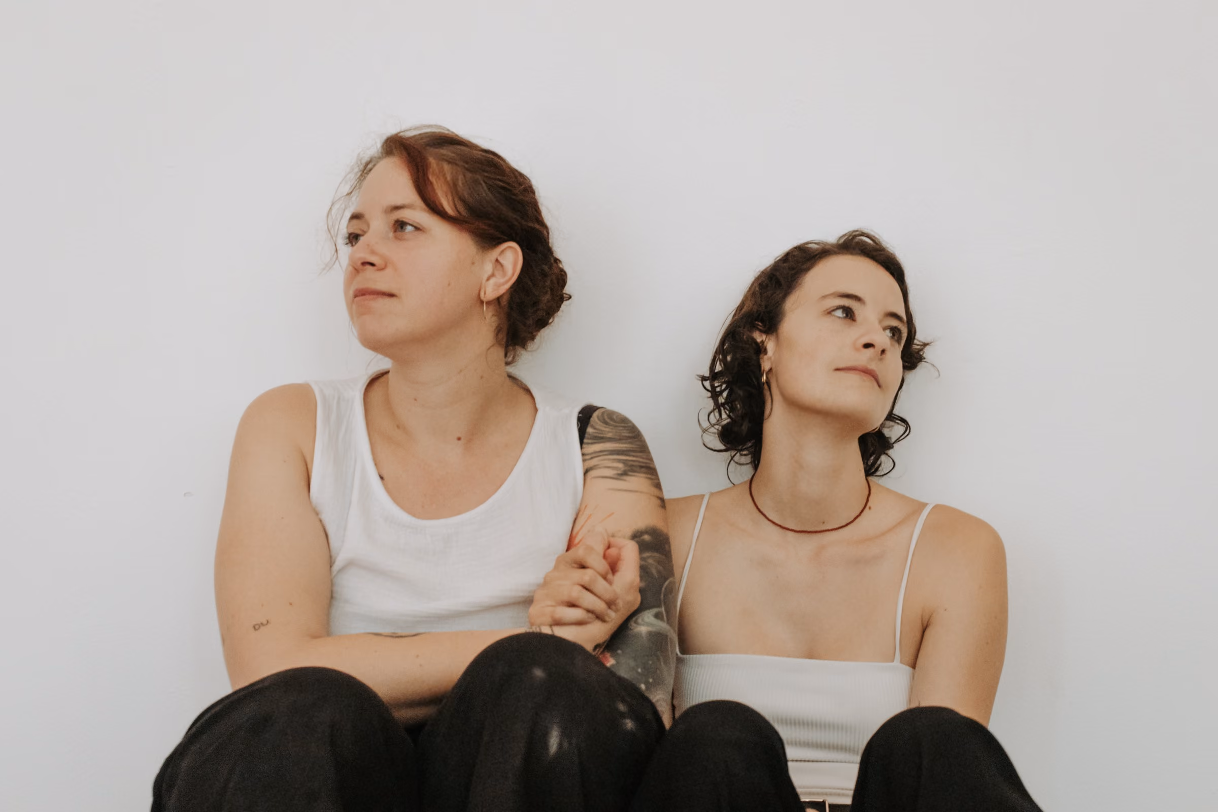 Karo und Pia sitzen nebeneinander an einer hellen Wand, die Blicke nachdenklich im Raum