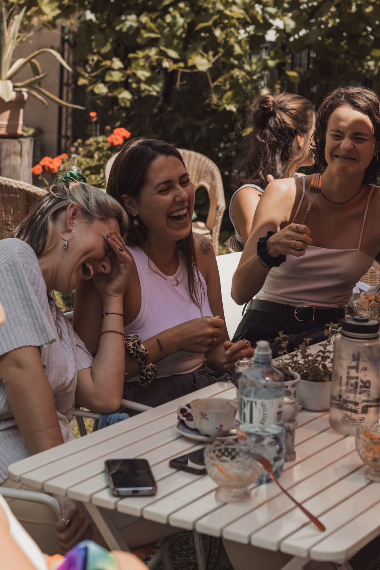 Teilnehmerinnen sitzen lachend an einem Tisch im Außenbereich des frei Yoga Café, Pflanzen und Geranien ringsum