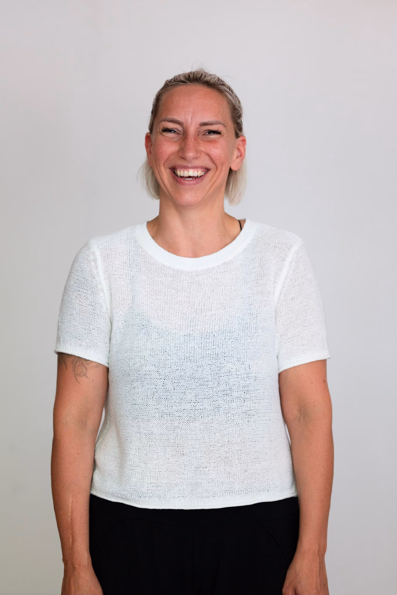 Portrait aus dem Kollektiv, lachend im weißen Strickshirt vor heller Wand